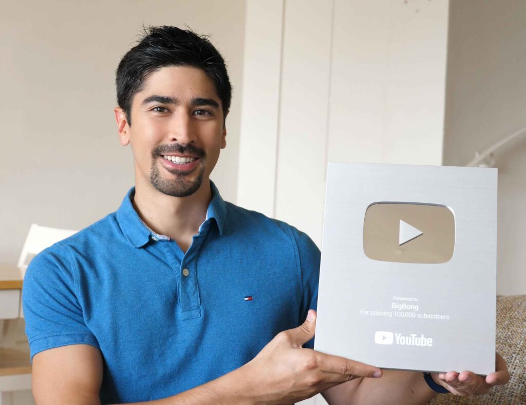 BigBong, online content creator, smiles in a blue polo shirt, proudly holding the YouTube Silver Award for reaching 100,000 subscribers.