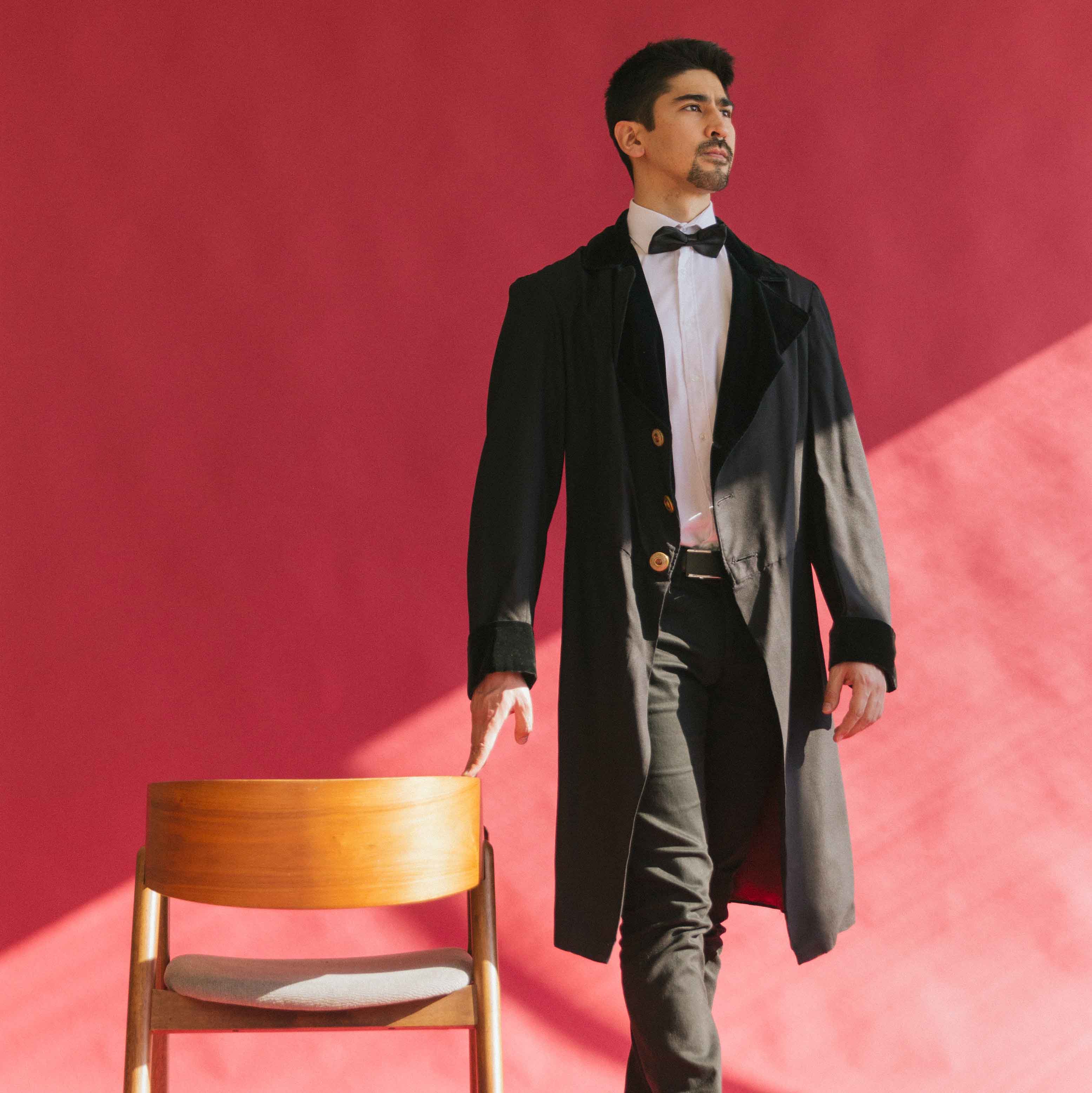 Model BigBong donning a bow-tie and a traditional long coat, striking a pose beside a chair with a vibrant red background – a stylish and sophisticated image.