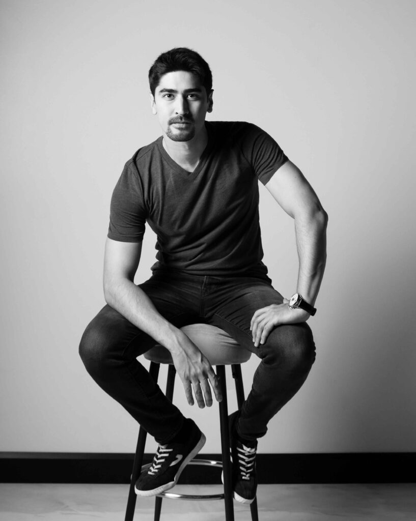 Black and white portrait of Model BigBong seated on a chair, one hand resting on his lap, with a direct gaze into the camera