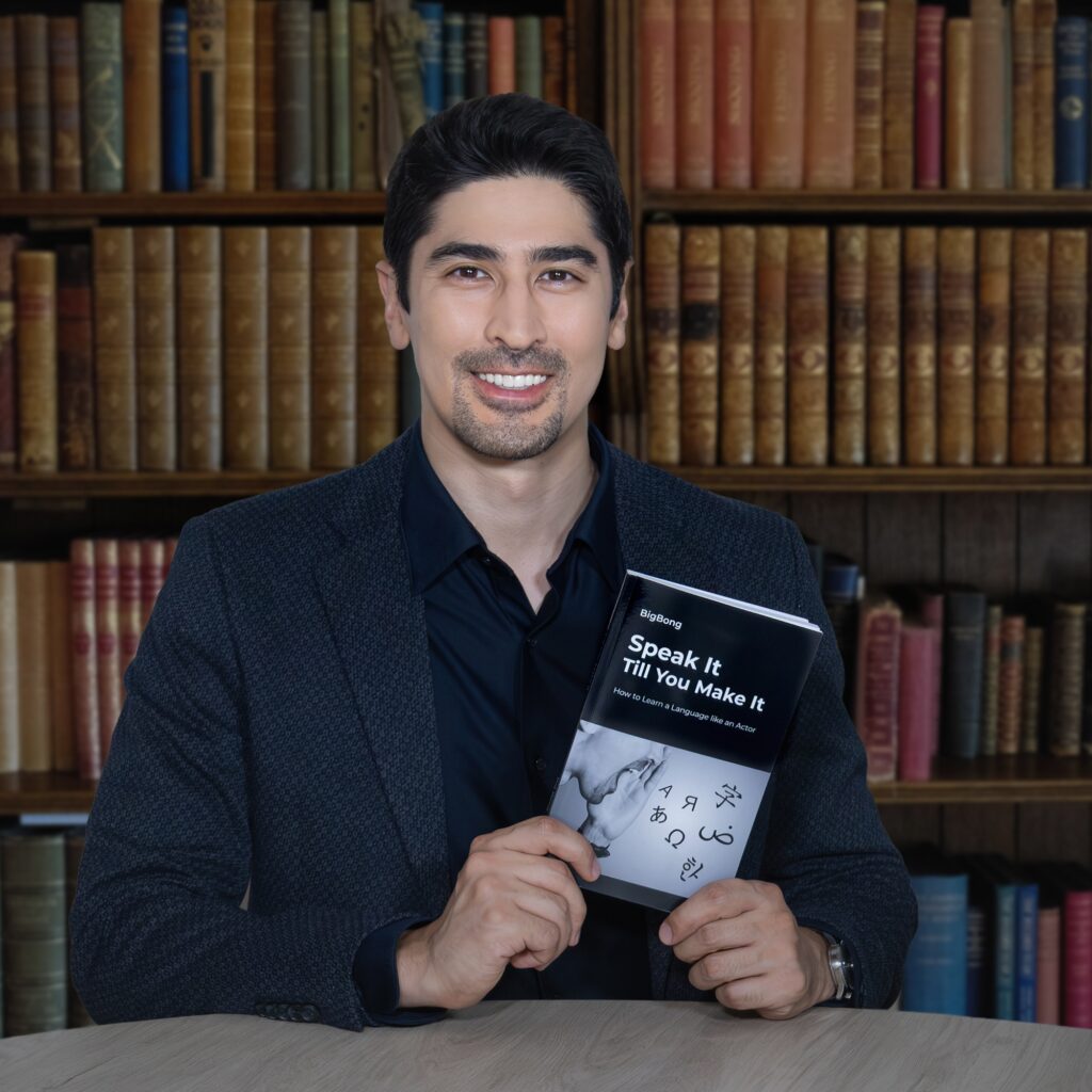 BigBong smiling and holding his book "Speak It Till You Make It," standing in front of a bookshelf in an old library.